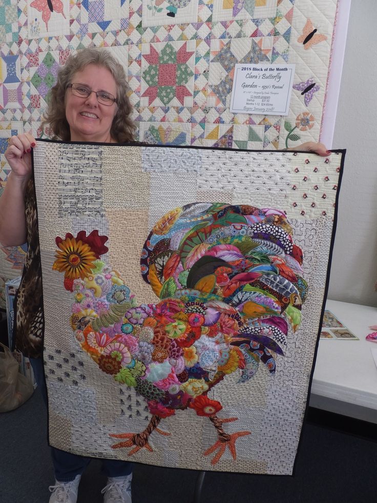 a woman holding up a quilt made to look like a rooster with flowers on it