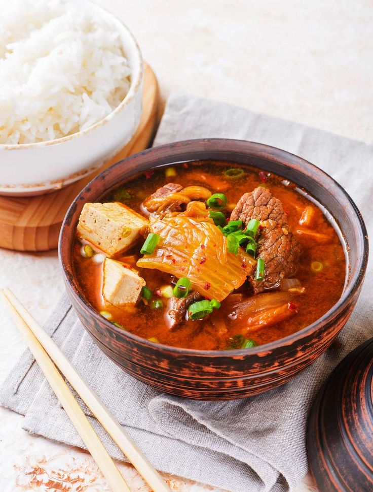 a bowl filled with stew next to rice and chopsticks on a table top