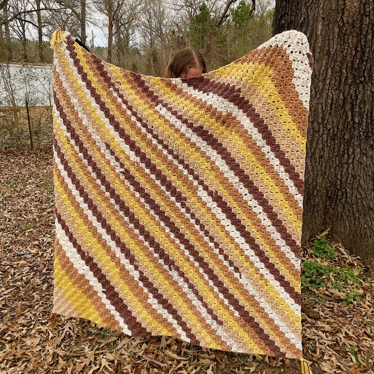 a woman holding up a crocheted blanket in front of a tree with leaves on the ground