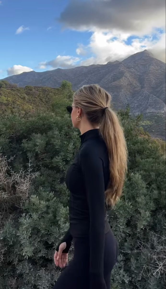 a woman standing on top of a hill next to trees and bushes with mountains in the background