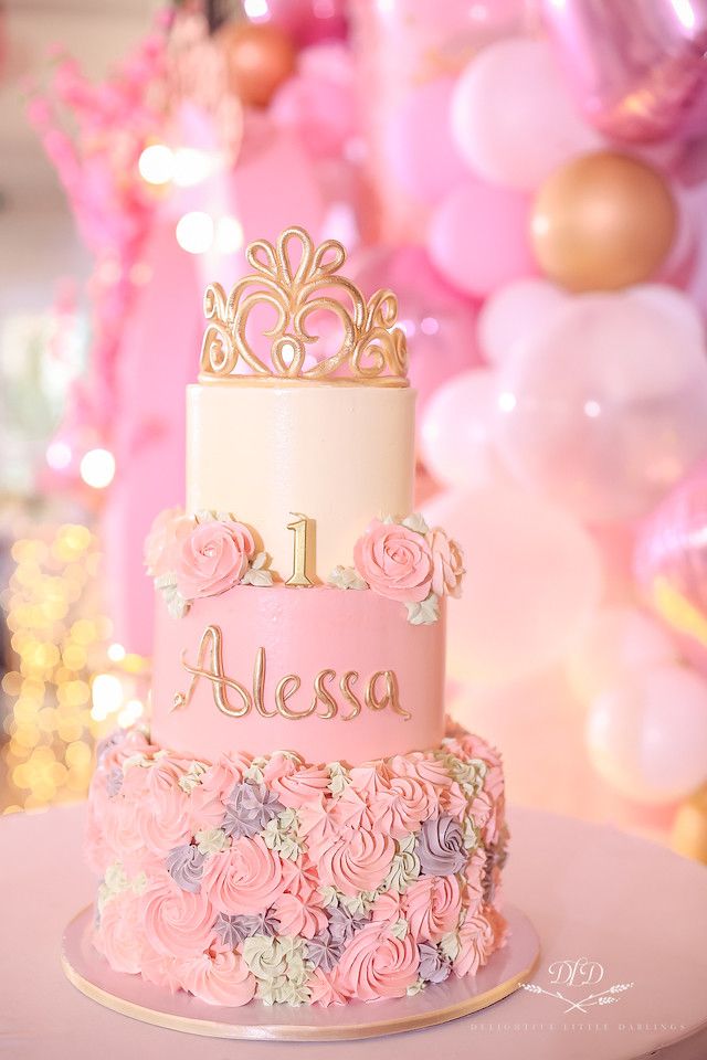 a pink and gold birthday cake with flowers on the bottom, sitting on a table in front of balloons