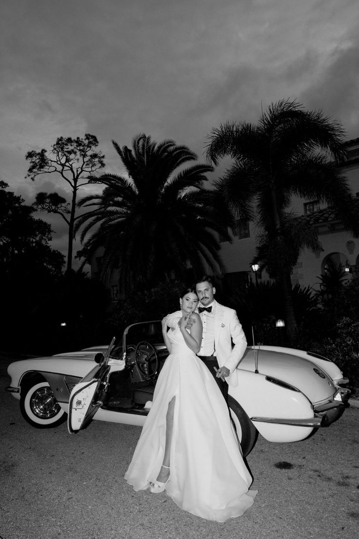 a bride and groom standing in front of a vintage car at night with palm trees behind them