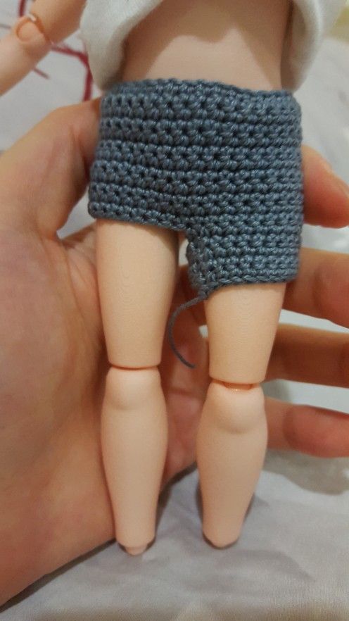 a hand holding a small doll in a knitted blue shorts and white shirt on top of a bed