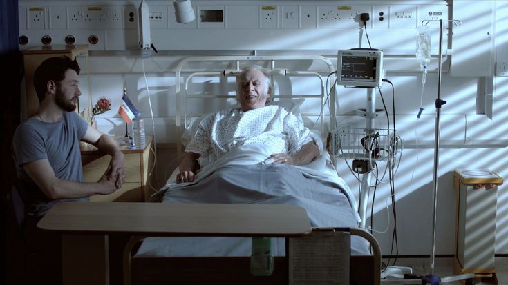 an old man in a hospital bed talking to a young man who is sitting next to him