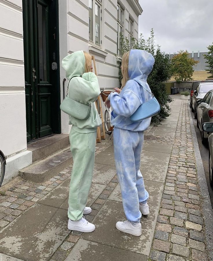 two people dressed in pastel clothing standing on the sidewalk next to parked cars and buildings