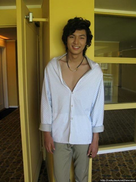 a young man standing in front of a yellow wall with his shirt open and wearing khaki pants