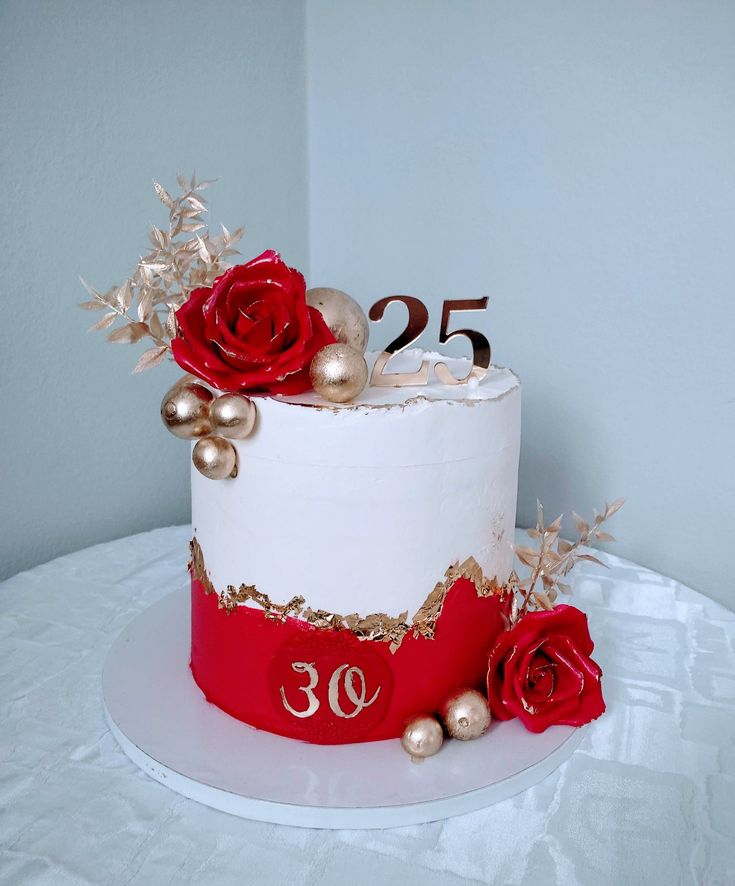 a white and red birthday cake with gold decorations on the top is decorated with roses