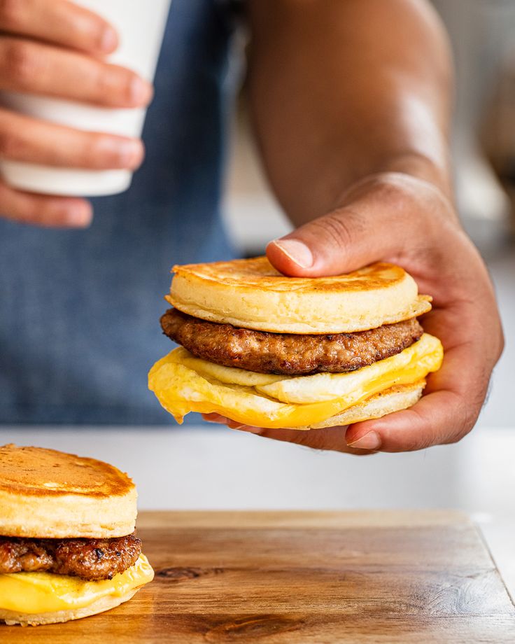 a person holding up a stack of pancakes with eggs and sausage on them, while another holds a cup of coffee in the background