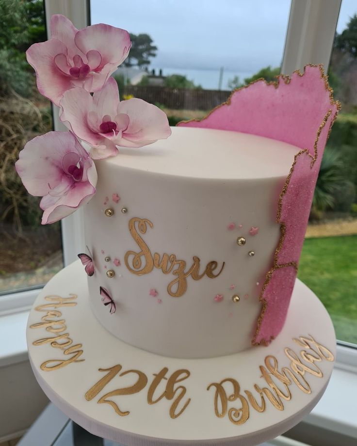 a white cake with pink flowers and gold writing on it, sitting in front of a window