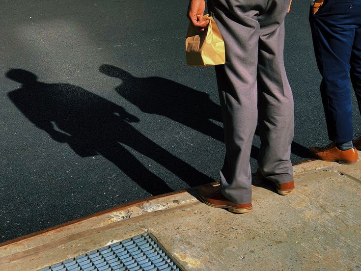 two people standing on the side walk with their shadows on the ground and one person holding a bag