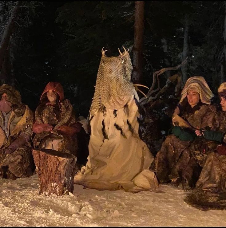 several people dressed up in costumes sitting on the ground with snow around them and trees behind them