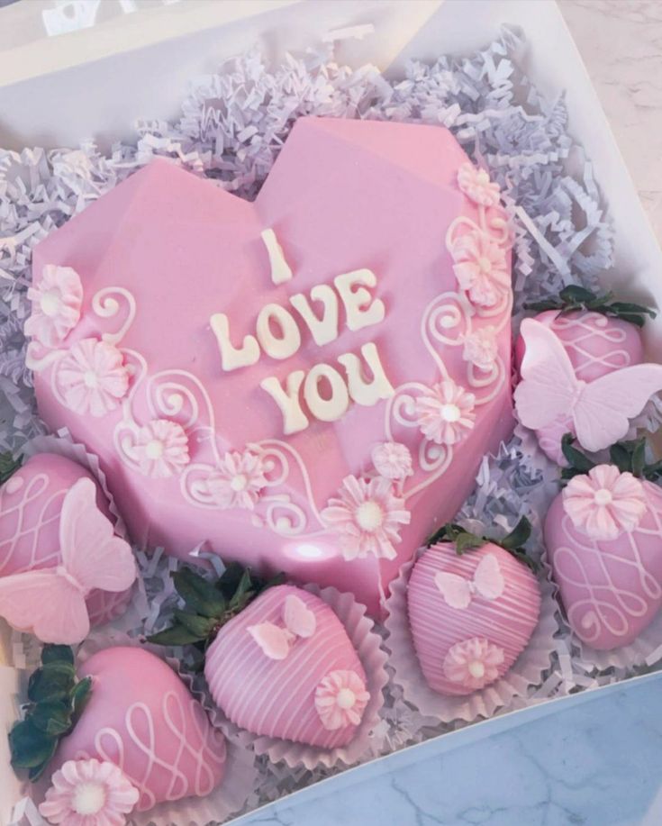 a pink heart shaped cake in a box with chocolates and flowers on the side