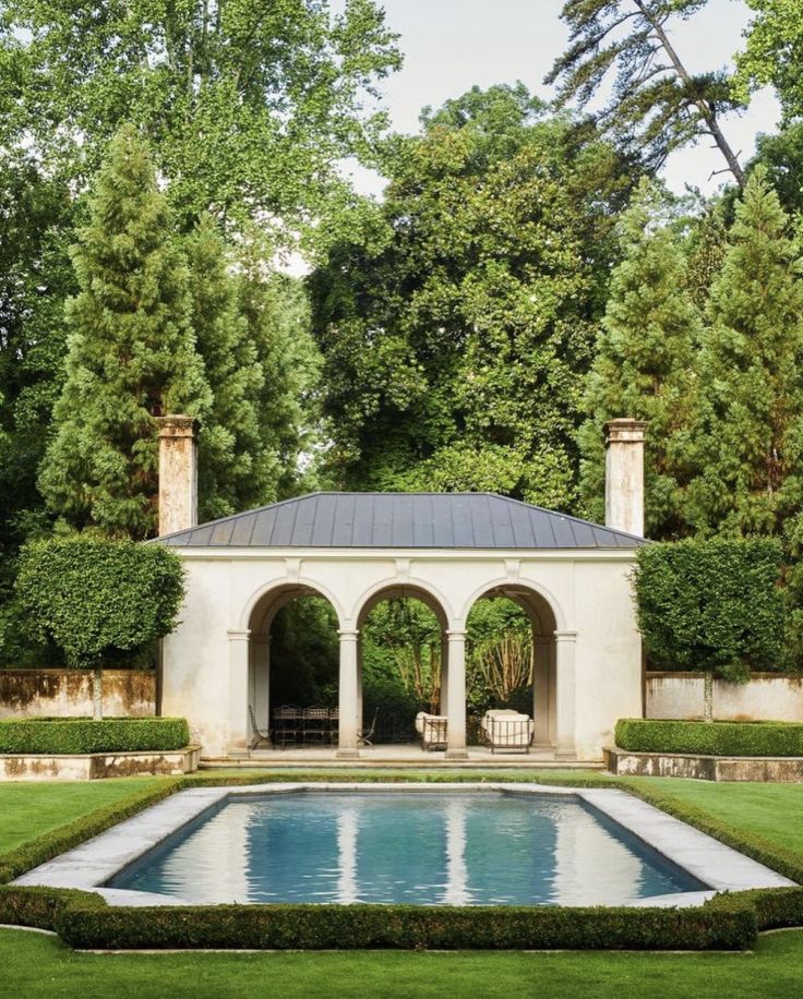 an outdoor pool surrounded by hedges and trees in the middle of a garden with a gazebo