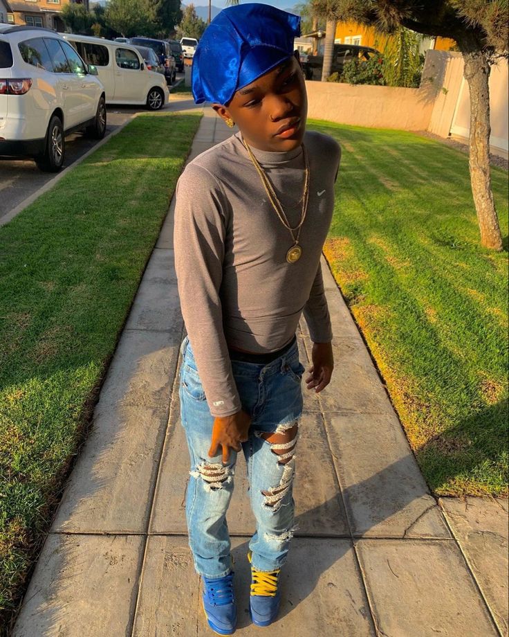 a young boy in ripped jeans and a blue hat standing on the sidewalk with his hands behind his head