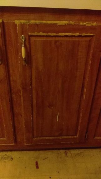 a wooden cabinet with peeling paint on the door and bottom panel, next to a toilet paper dispenser