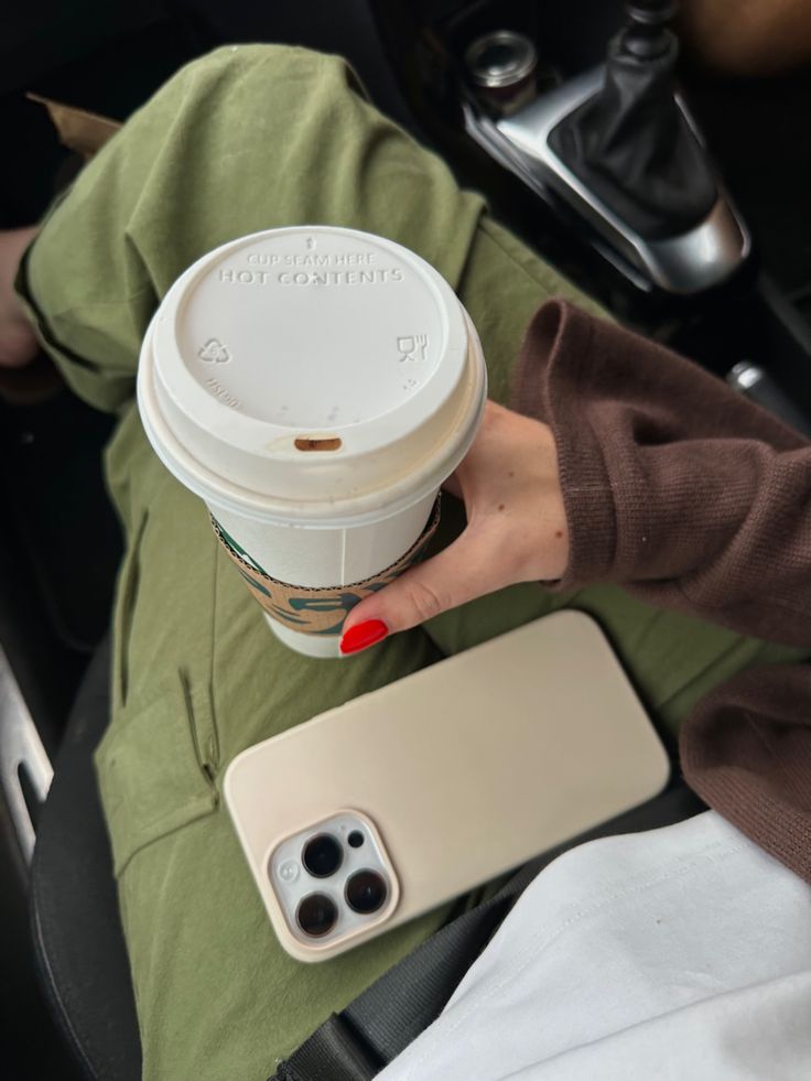 a woman holding a cup of coffee while sitting in a car with her cell phone