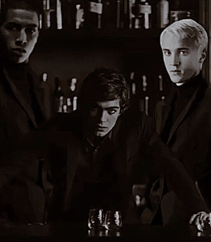 three men sitting at a bar in front of bottles and glasses, one wearing a black suit