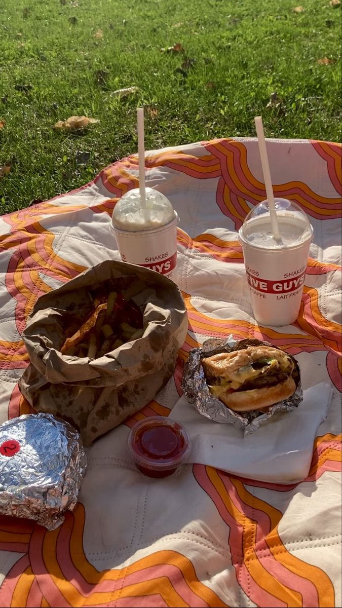 a picnic blanket with food on it and two cups of milkshakes in the background