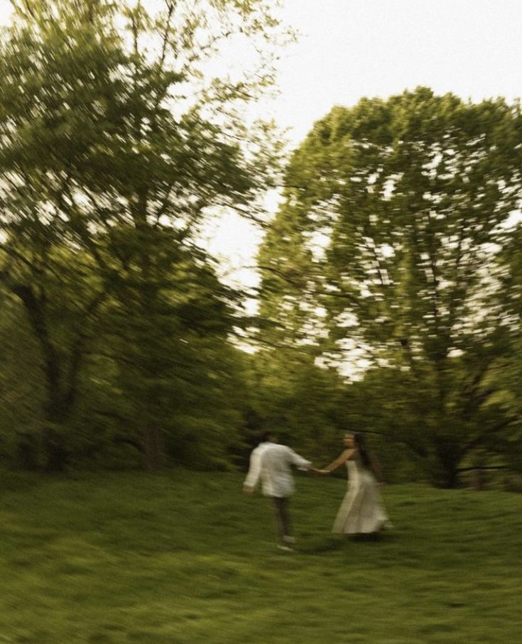 a man and woman are walking in the grass holding hands with trees in the background