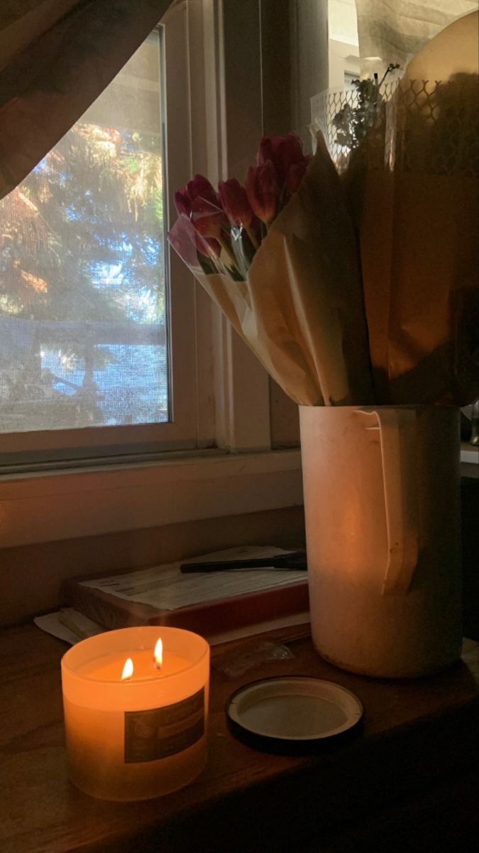 a lit candle and some flowers in a vase on a window sill by a window