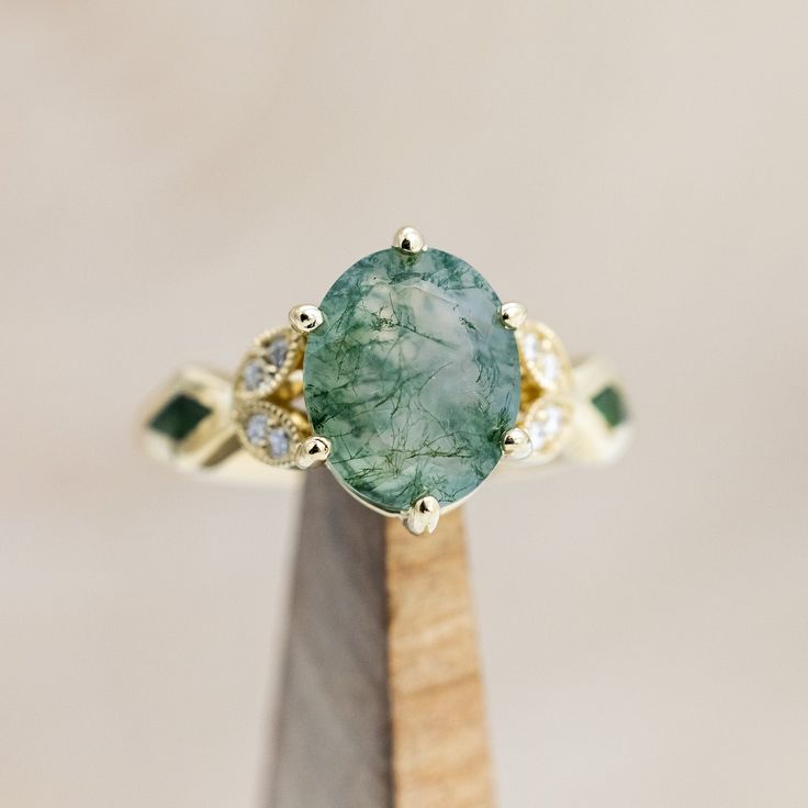 a close up of a ring with a green stone and diamonds on the top, sitting on a wooden stand