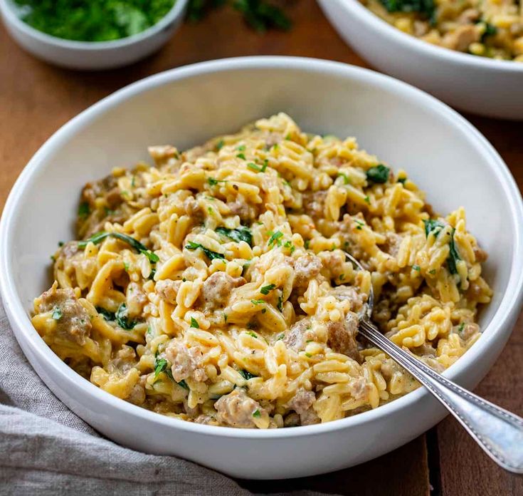 two white bowls filled with pasta and spinach on top of a wooden table next to silverware