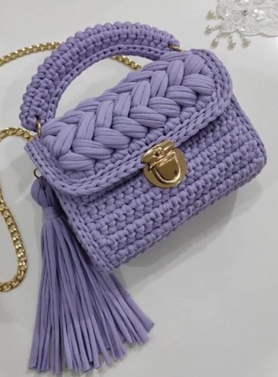 a purple handbag with a tasselled handle on a white surface next to a gold chain