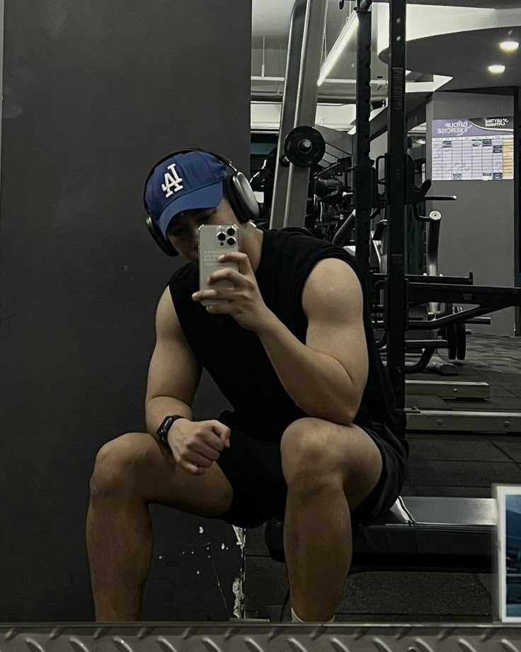 a man sitting on top of a bench in a gym holding a cell phone up to his face