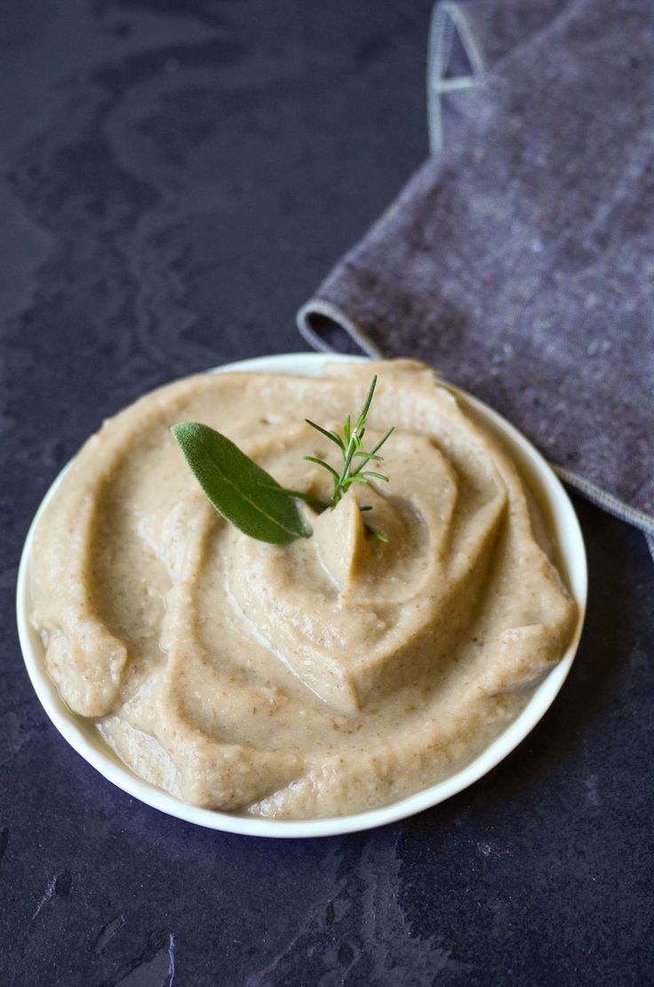 a white plate topped with hummus next to a gray napkin and green sprig