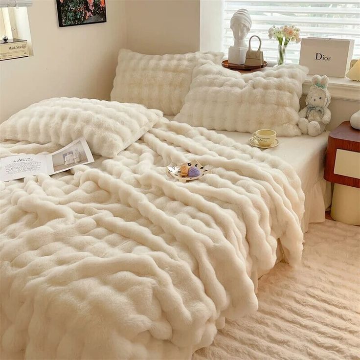 a bed covered in white fluffy blankets next to a window with a teddy bear on it