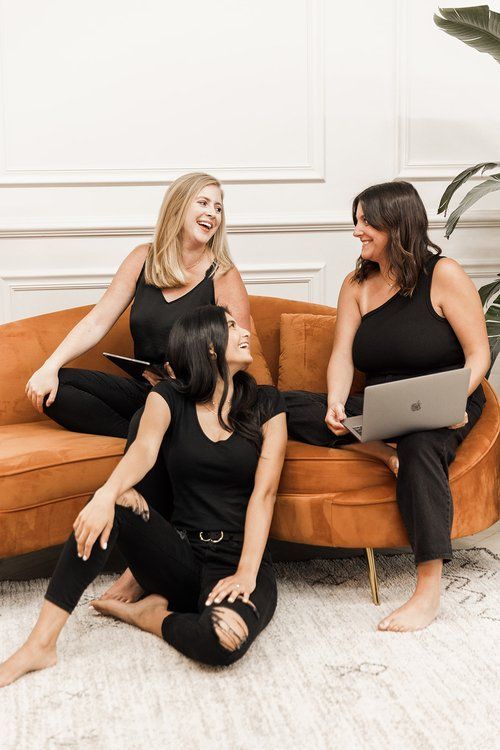 three women sitting on a couch laughing and looking at each other with laptops in their hands