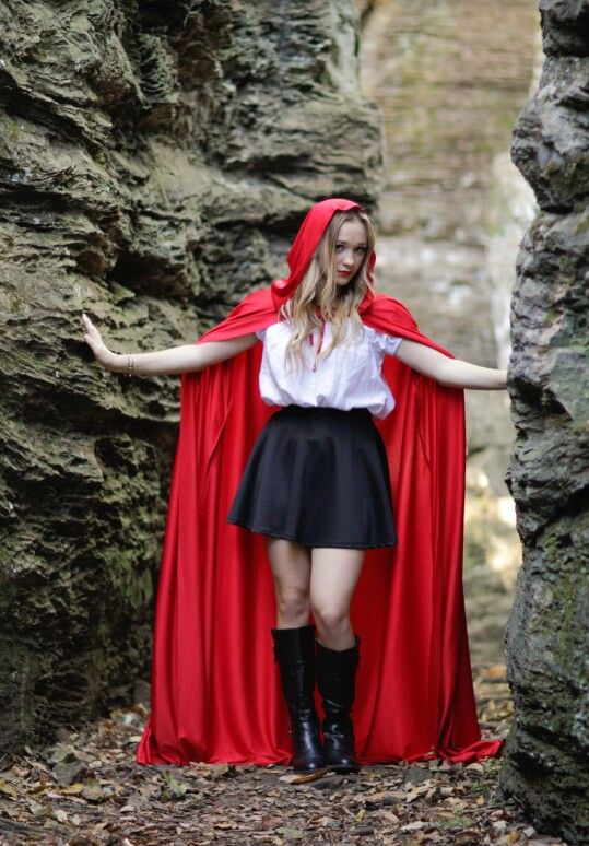 a woman in a red cape and black skirt is standing by some rocks with her arms outstretched