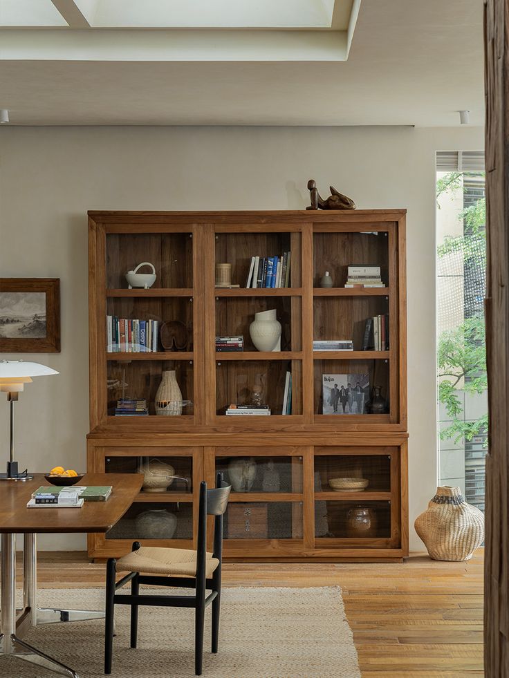 a wooden bookcase with glass doors in a living room next to a table and chair