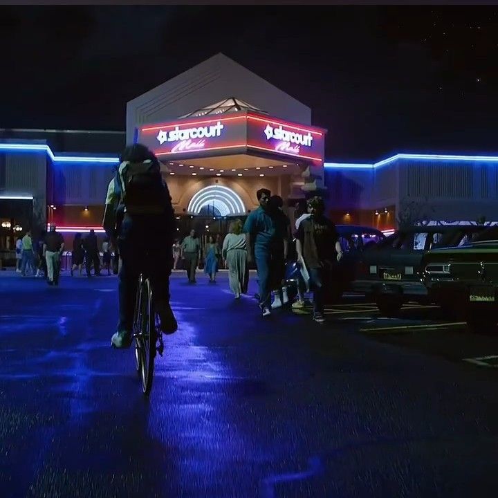 people are walking and riding bikes in front of a building at night with neon lights