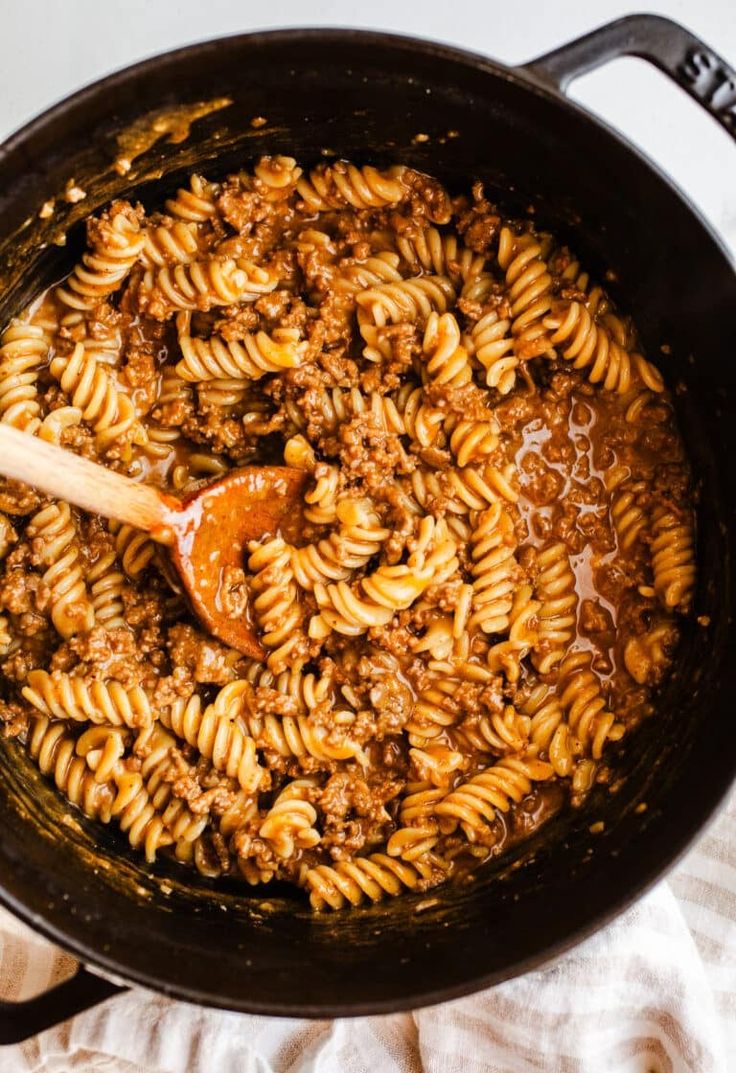 a skillet filled with pasta and sauce on top of a white cloth next to a wooden spoon