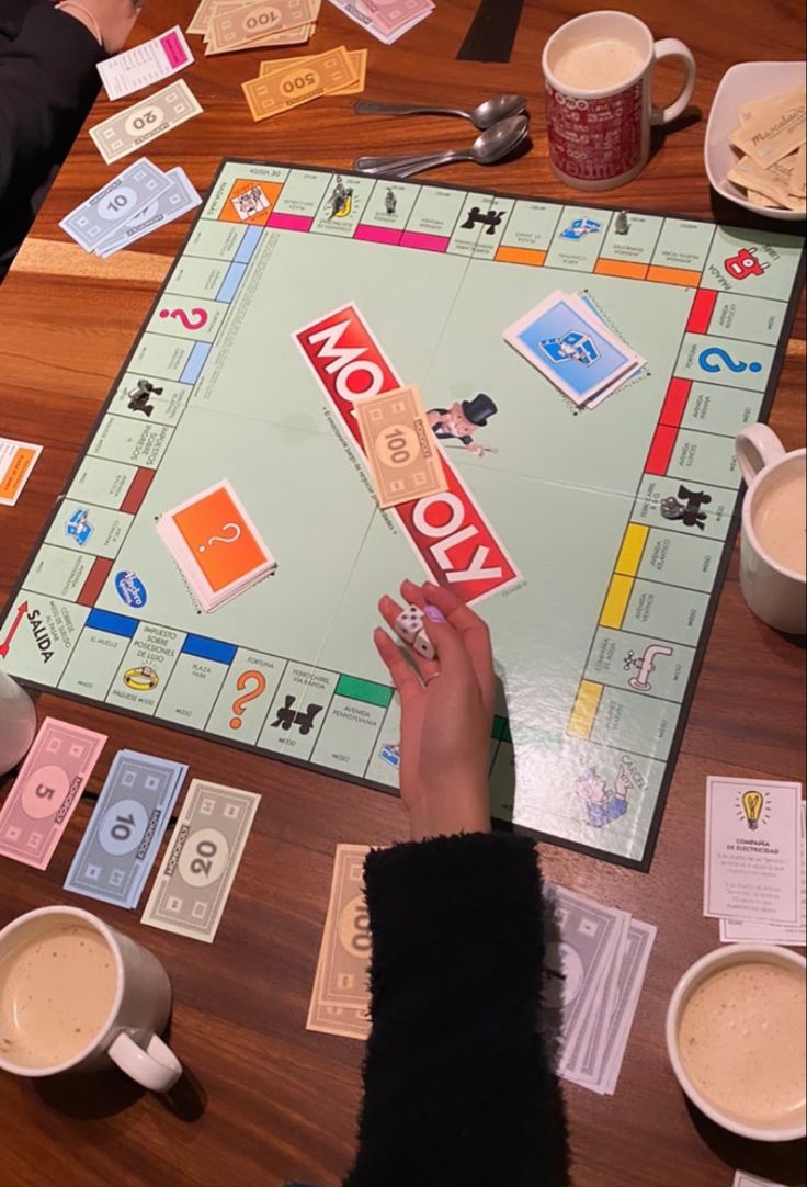 two people playing monopoly on a wooden table with cups of coffee and spoons nearby