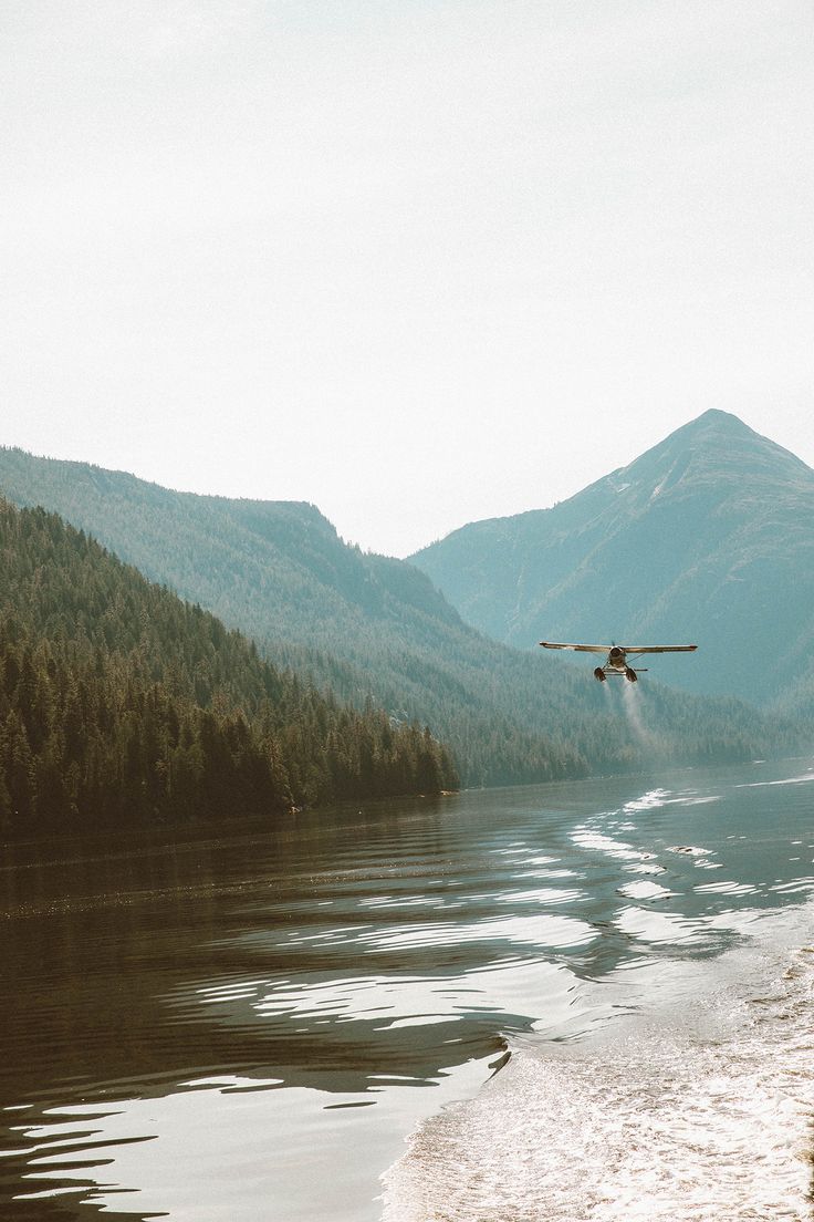 an airplane is flying over the water in front of some mountains and trees on the shore