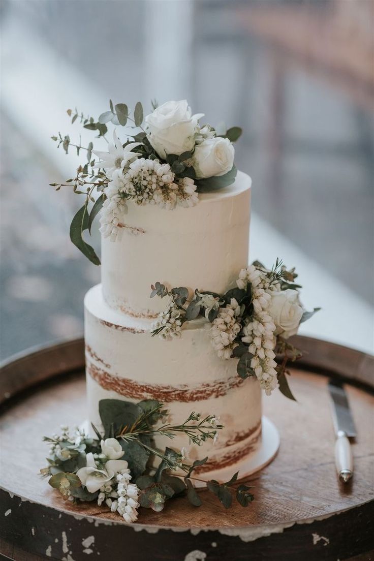 a three tiered cake with flowers and greenery sits on top of a wooden table