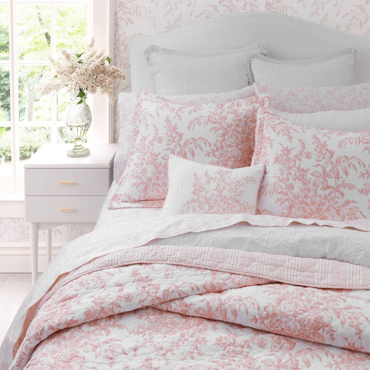 a bed with pink and white comforters in a bedroom next to a window filled with flowers