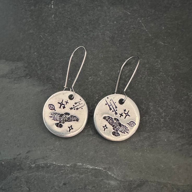 two silver earrings with designs on them sitting on top of a black surface and one has a white disc