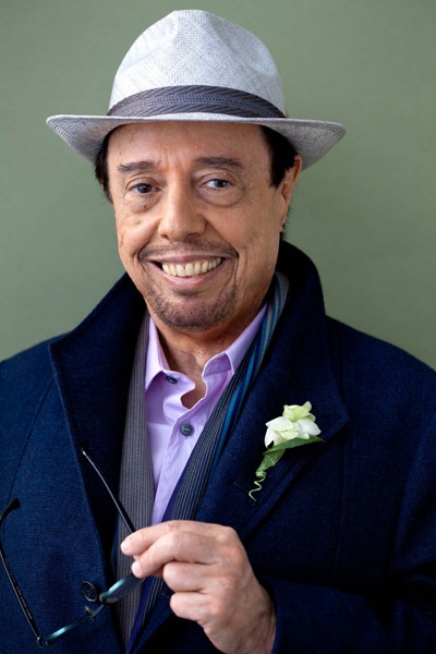 a man in a suit and hat smiles while holding a pair of scissors to his left