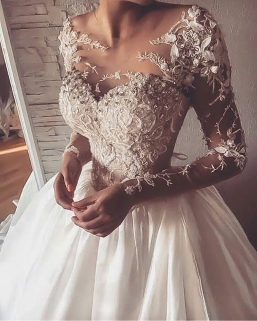 a woman wearing a wedding dress with long sleeves and flowers on the shoulder, standing in front of a mirror