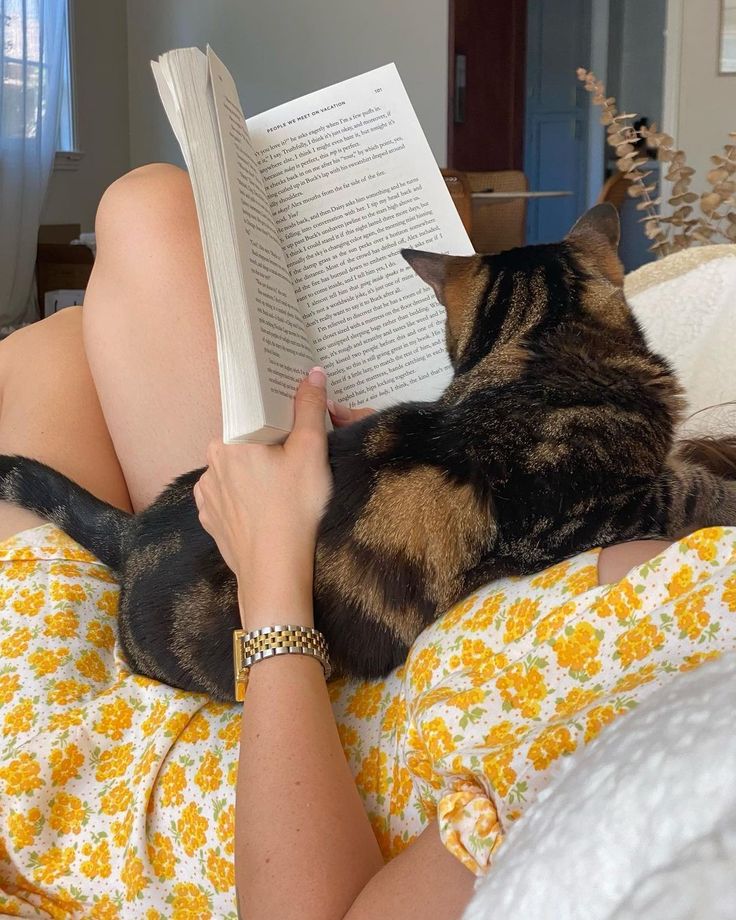 a woman reading a book while laying on her stomach with a cat in her lap