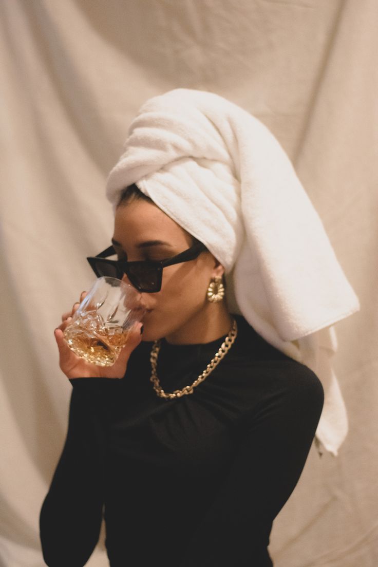 a woman with a towel on her head drinking from a wine glass in front of a white backdrop