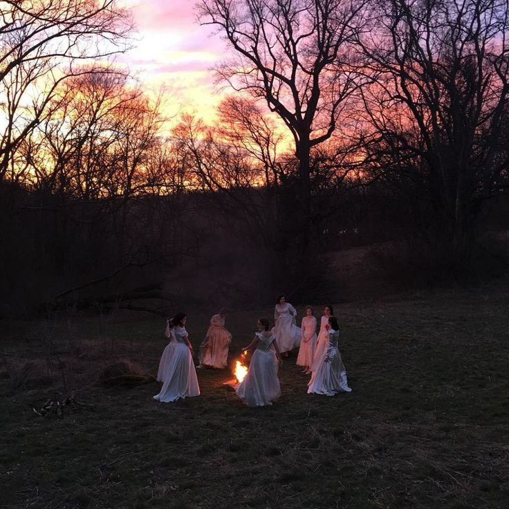 a group of women in white dresses standing around a fire pit at sunset with trees in the background