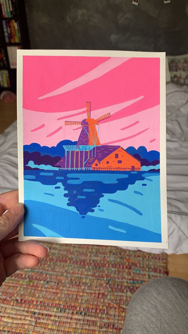 a person holding up a card with a painting of a windmill on the water in front of them