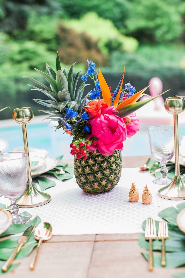 a pineapple centerpiece with colorful flowers and greenery sits on a table next to silverware