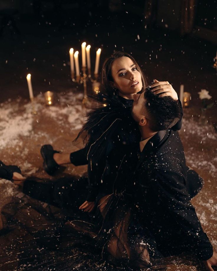 a woman sitting on the ground next to a man in black clothes with candles behind her