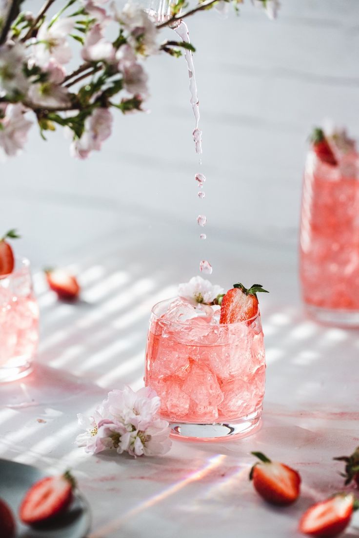 two glasses filled with ice and strawberries on top of a white table next to flowers