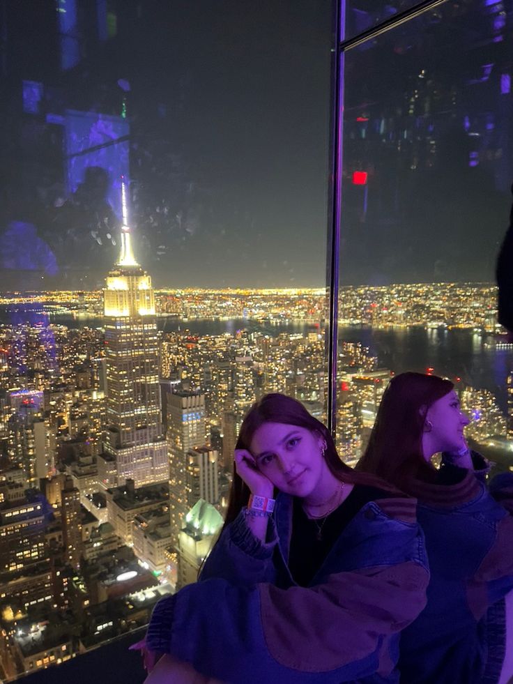 two people standing in front of a window at the top of a tall building with city lights behind them